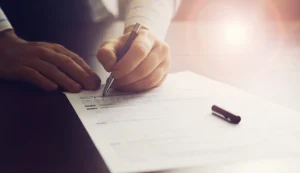 A person is holding a pen and filling out a printed form on a table, with sunlight shining in the background.