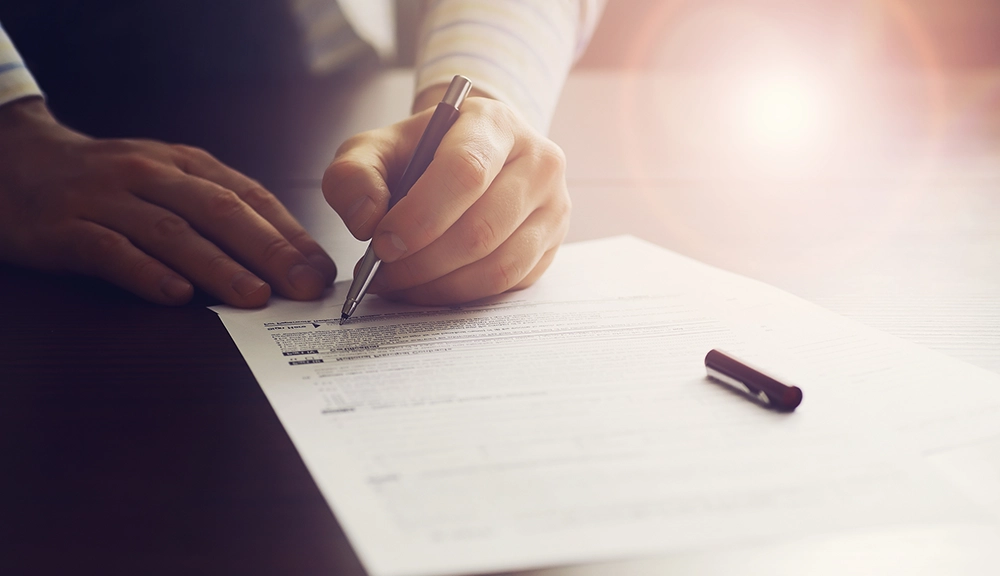 A person is holding a pen and filling out a printed form on a table, with sunlight shining in the background.