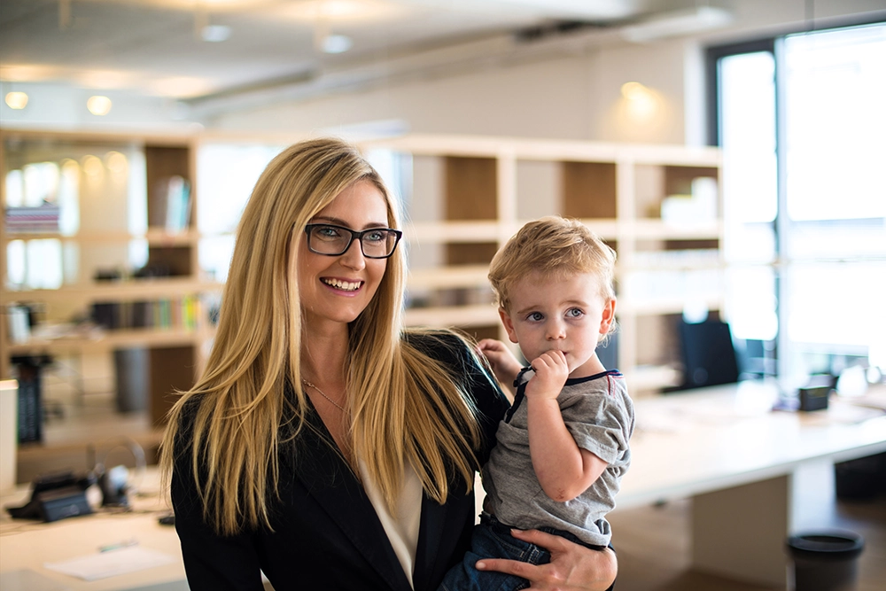A woman wearing glasses and a blazer stands in an office, smiling while holding a young boy who has his hand near his mouth.