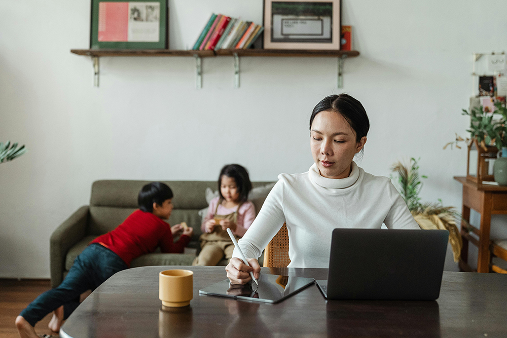 A woman works at a table with a laptop and tablet, while two children play on a sofa in the background.