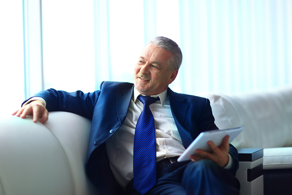 A man in a blue suit sits on a white couch, smiling and holding a tablet with one hand. Bright, natural light fills the room.