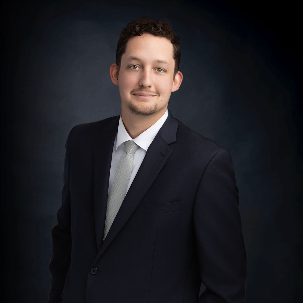 A man wearing a dark suit, white shirt, and light tie poses against a dark, gradient background.