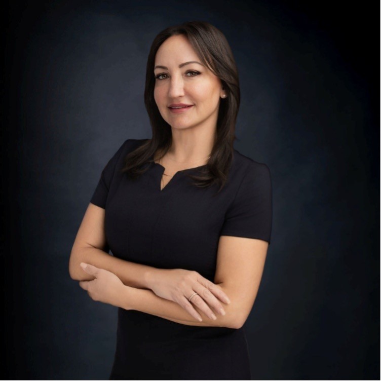 Woman with long brown hair wearing a dark short-sleeve dress stands against a dark background with arms crossed, looking at the camera and smiling slightly.