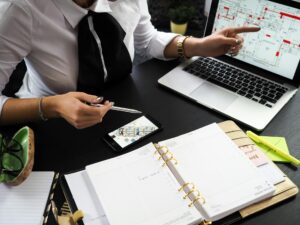 Person in business attire sits at a desk with an open planner, a smartphone, papers, and a laptop displaying architectural plans, pointing at the laptop screen.