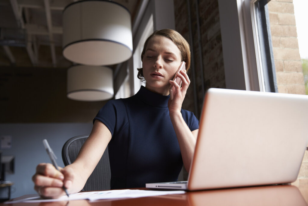 A person sits at a desk with a laptop, talking on a phone and writing on paper, inside an office with large windows and modern lighting.