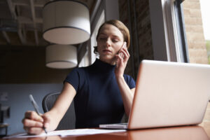 A person sits at a desk with a laptop, talking on a phone and writing on paper, inside an office with large windows and modern lighting.