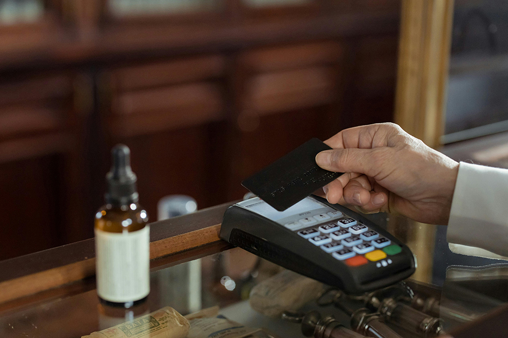 A person holds a black credit card above a payment terminal on a glass counter, ready for a contactless transaction; a dropper bottle and cash are also visible.