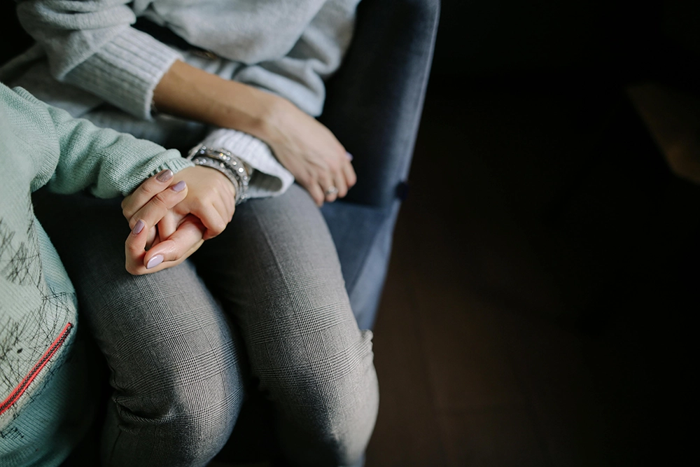 Two people sit side by side on a chair, one wearing a grey sweater and pants, holding hands with another person in a green sweater.