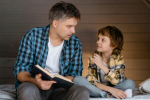 An adult and a child sit on a bed together; the adult holds an open book while the child looks at him and listens.