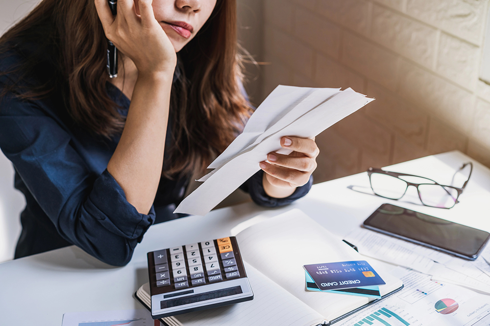 A woman sits at a desk talking on the phone, holding bills, with a calculator, credit cards, notebook, and documents spread out in front of her.