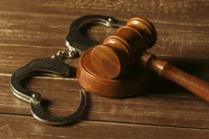 A wooden judge's gavel rests next to a pair of open metal handcuffs on a wooden table.
