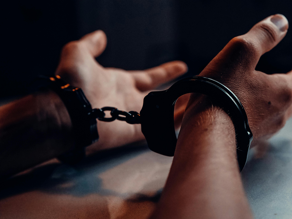 Close-up of a person's hands restrained with black handcuffs on a table, with light and shadow emphasizing the wrists and chain.