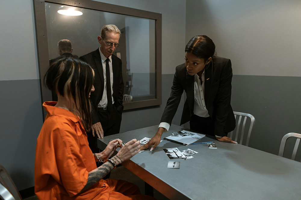 A person in an orange jumpsuit sits at a table with two people in suits; one is showing them photos during an interrogation.