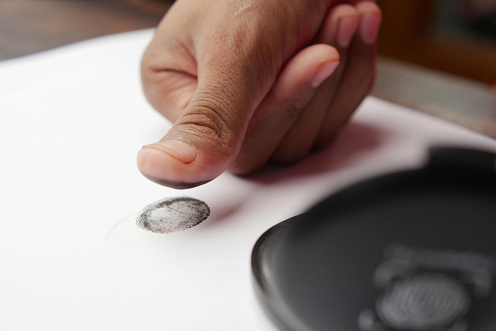 A person presses their inked thumb onto white paper, leaving a clear fingerprint beside an ink pad.
