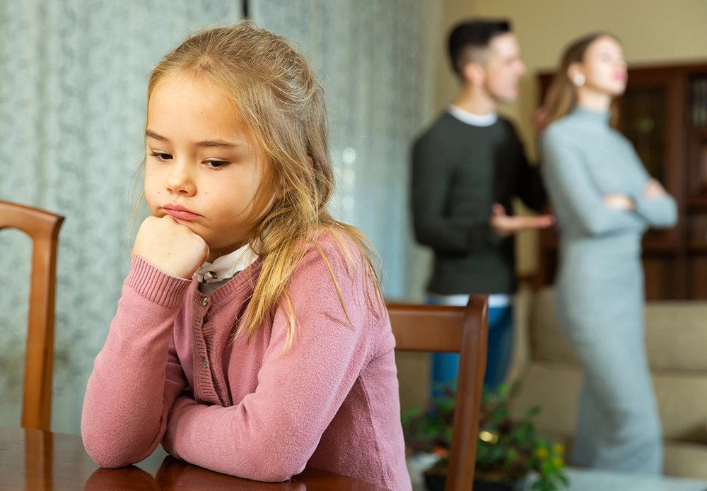 A young girl sits at a table looking sad, while a man and a woman argue in the background, both facing away from each other.