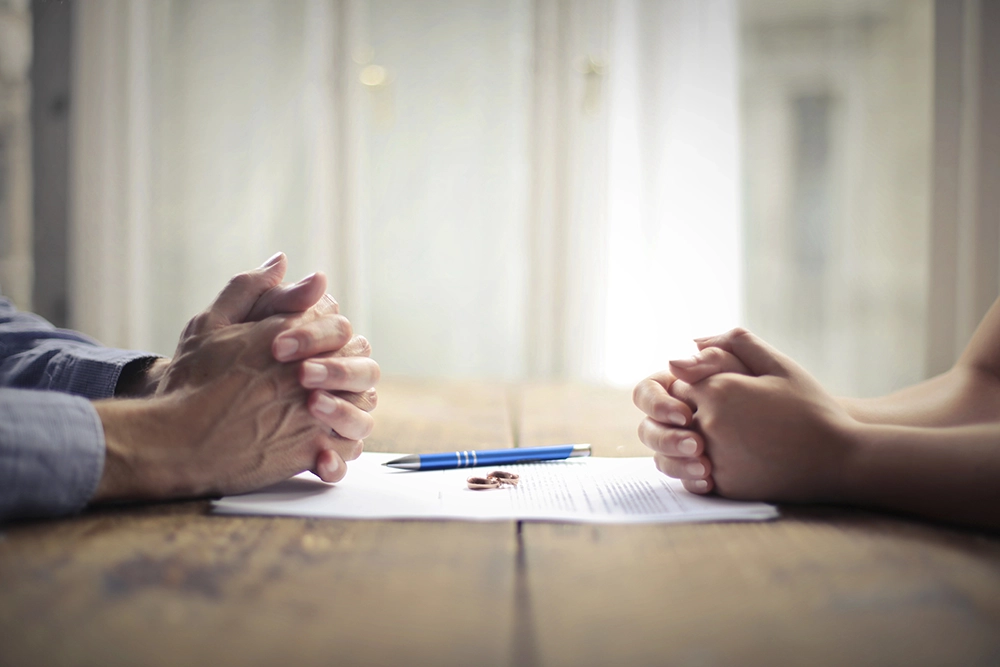 Two people sit across from each other at a table with hands clasped, a pen, and a document between them.
