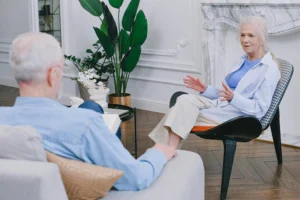An older woman sits in a chair speaking to an older man sitting on a sofa in a well-decorated room with plants and a marble fireplace.