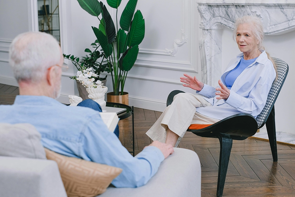 An older woman sits in a chair speaking to an older man sitting on a sofa in a well-decorated room with plants and a marble fireplace.