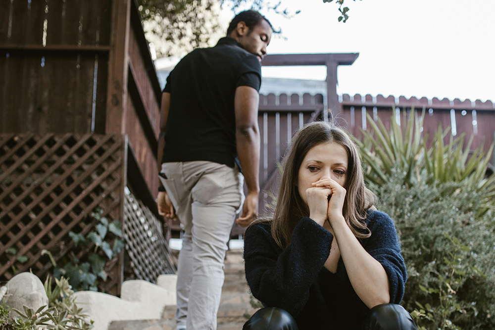 A woman sits on outdoor steps looking pensive, while a man walks away up the steps, glancing back at her.