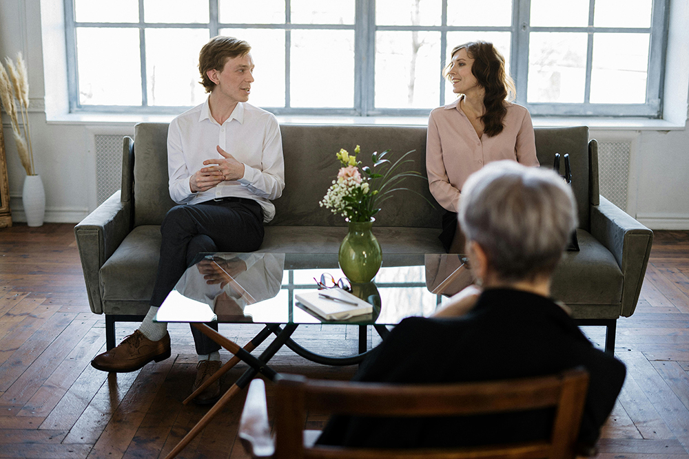 A man and a woman sit on a sofa facing a therapist, with a vase of flowers and a notebook on the glass coffee table between them.