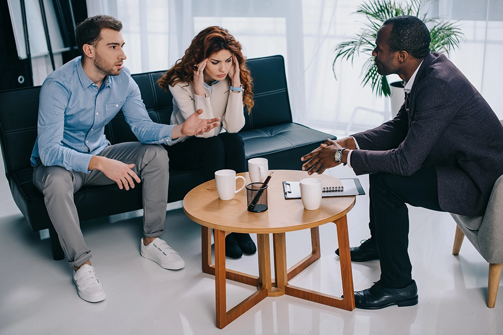 A couple sits on a couch speaking with a professional man across a coffee table with notebooks, hourglass, and mugs on it.