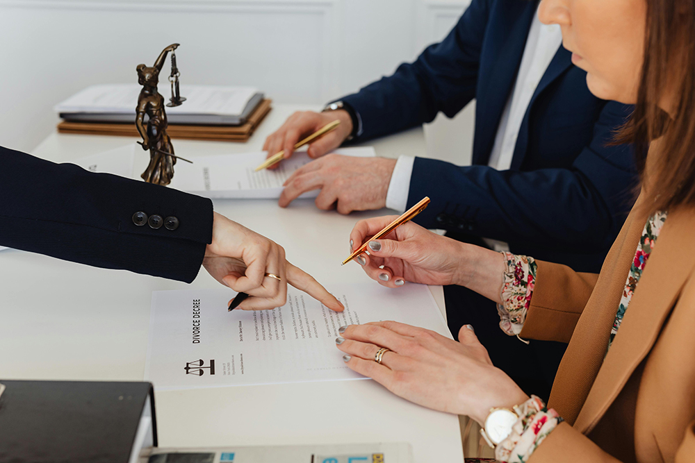 Three people sit at a desk reviewing and signing divorce papers, with one person pointing at a specific section on the document.