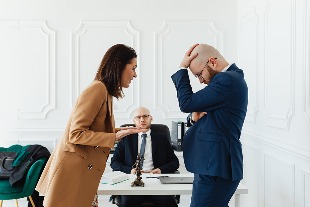 A woman in a tan blazer argues with a man in a blue suit, who looks distressed, while another man sits at a desk watching them.