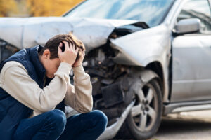 A person crouches with their head in their hands in front of a damaged car with a crumpled front end after an apparent accident.