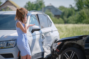 Woman standing by a damaged white SUV, talking on the phone after a car accident with another black car on a sunny day.