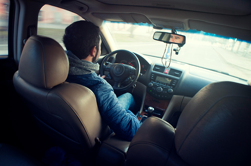 A person wearing a blue jacket drives a car, viewed from the back seat, with hands on the steering wheel and road visible through the windshield.