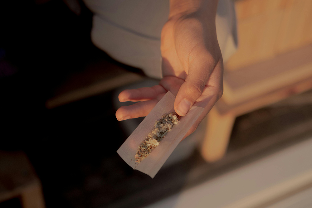 A hand holds a partially rolled cigarette or joint with loose tobacco or herbs visible inside the rolling paper.
