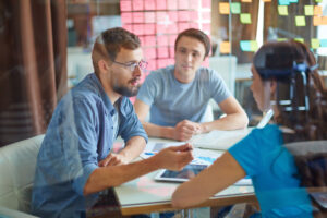 Three people sit at a desk in an office discussion, with documents and a tablet in front of them and a glass wall covered in colorful sticky notes in the background.