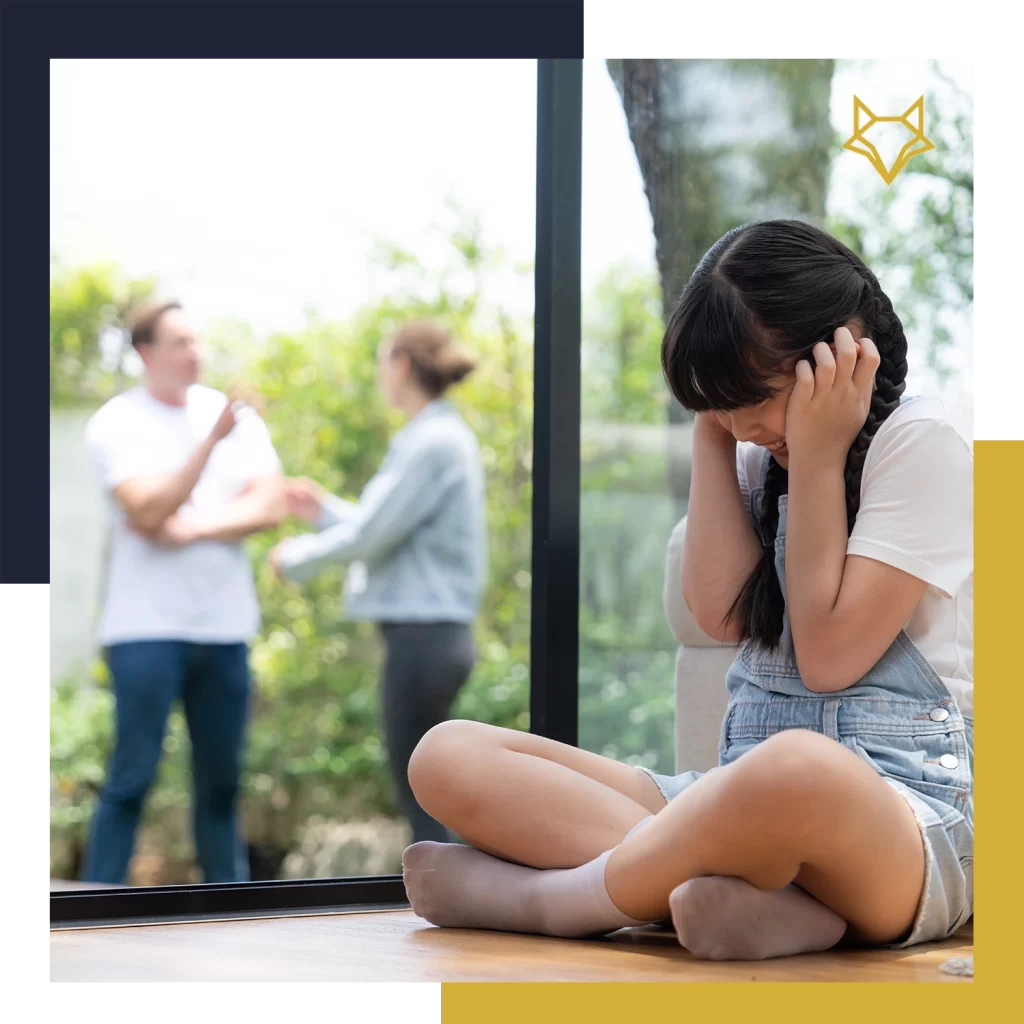 A young girl sits on the floor covering her ears while two adults argue in the background near a window.