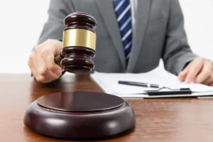 A person in a suit holds a gavel above a wooden sound block, with documents and a pen on the desk in the background.