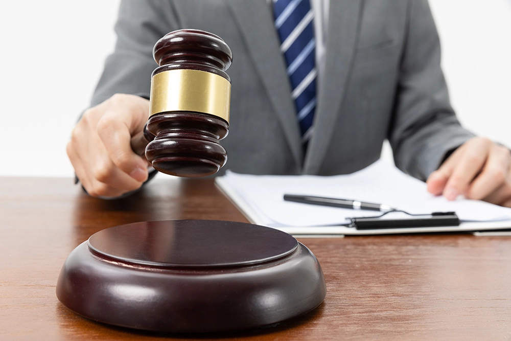 A person in a suit holds a gavel above a wooden sound block, with documents and a pen on the desk in the background.