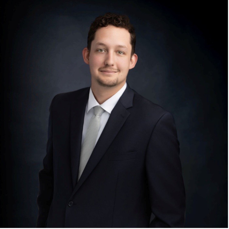 A man with short curly hair and a trimmed beard wearing a dark suit, white shirt, and light-colored tie poses against a dark, gradient background.