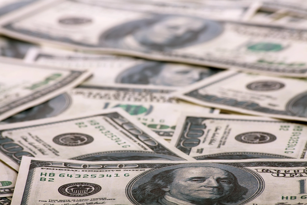 Several U.S. one hundred dollar bills are scattered in a pile, displaying Benjamin Franklin’s portrait and other recognizable security features.