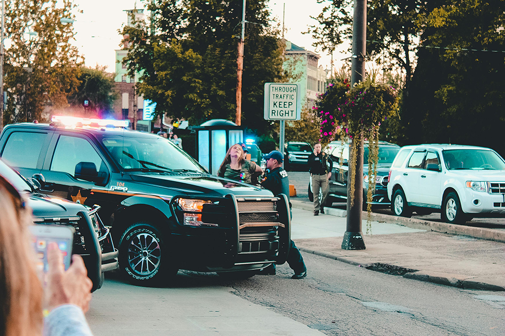 A police officer speaks to a woman near a black police SUV with flashing lights on a city street as bystanders look on.