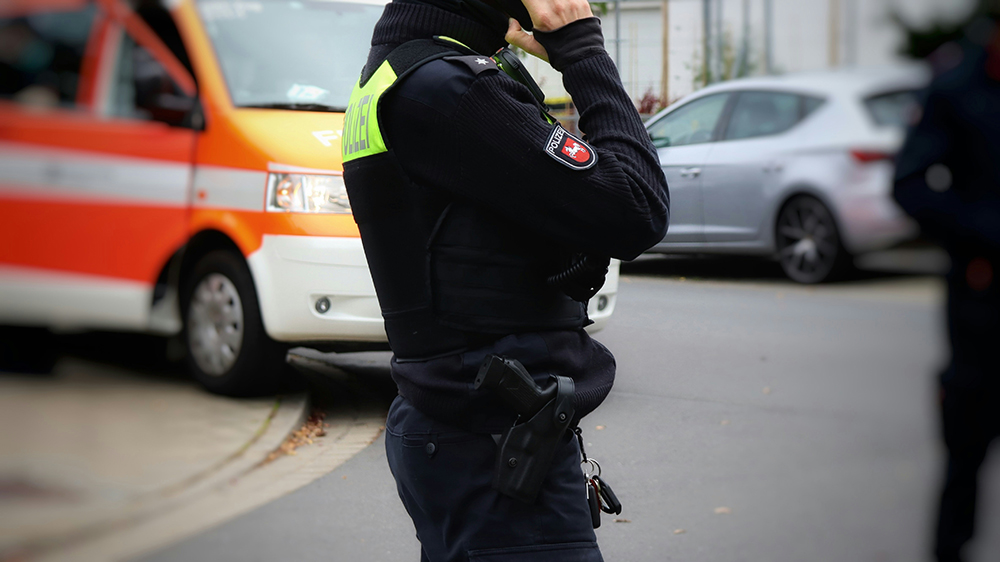 A police officer wearing a vest stands on a street, talking on a radio, with an emergency vehicle and cars in the background.