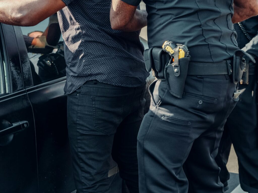 A police officer detains a person against a car, with the officer’s equipment and uniform clearly visible.